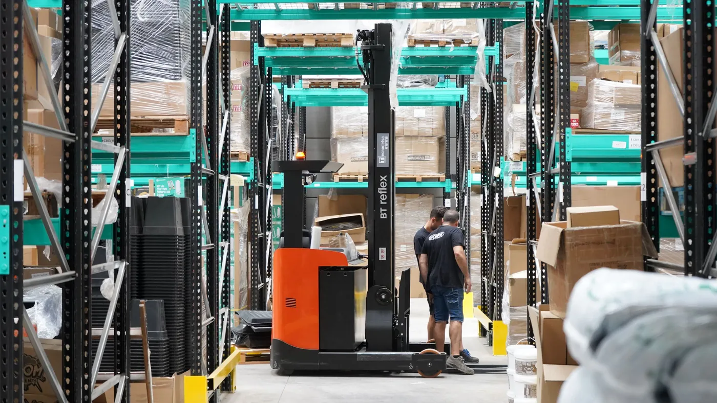 Two workers and a forklift between tall pallet racks in a distribution warehouse, representing business-to-business ordering, where AI agents have now completed live purchases without a sales rep.