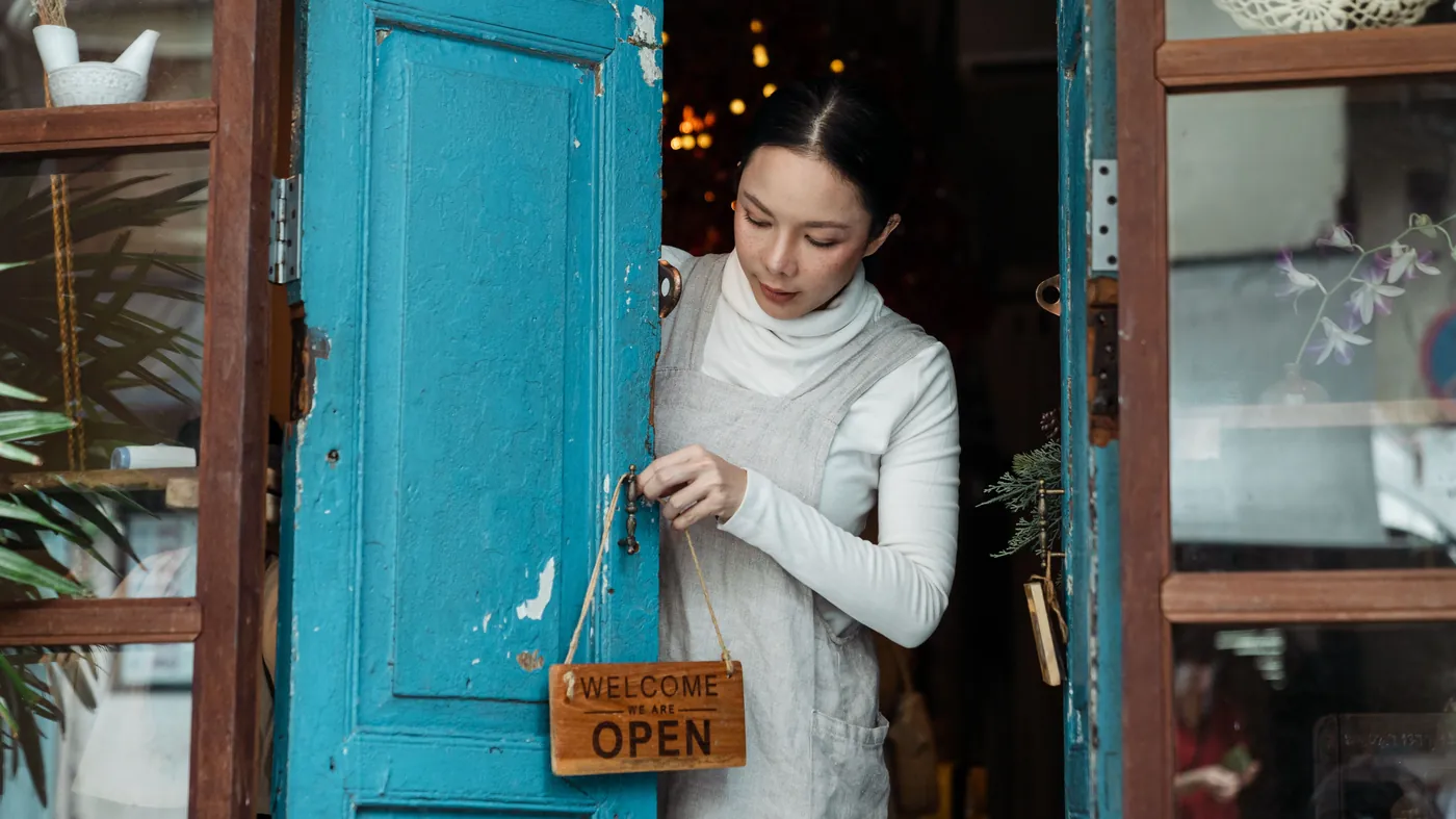 A shop owner turns a hand-lettered 'Welcome, we are open' sign on a weathered blue door, the physical version of a store signaling that it's ready for customers.