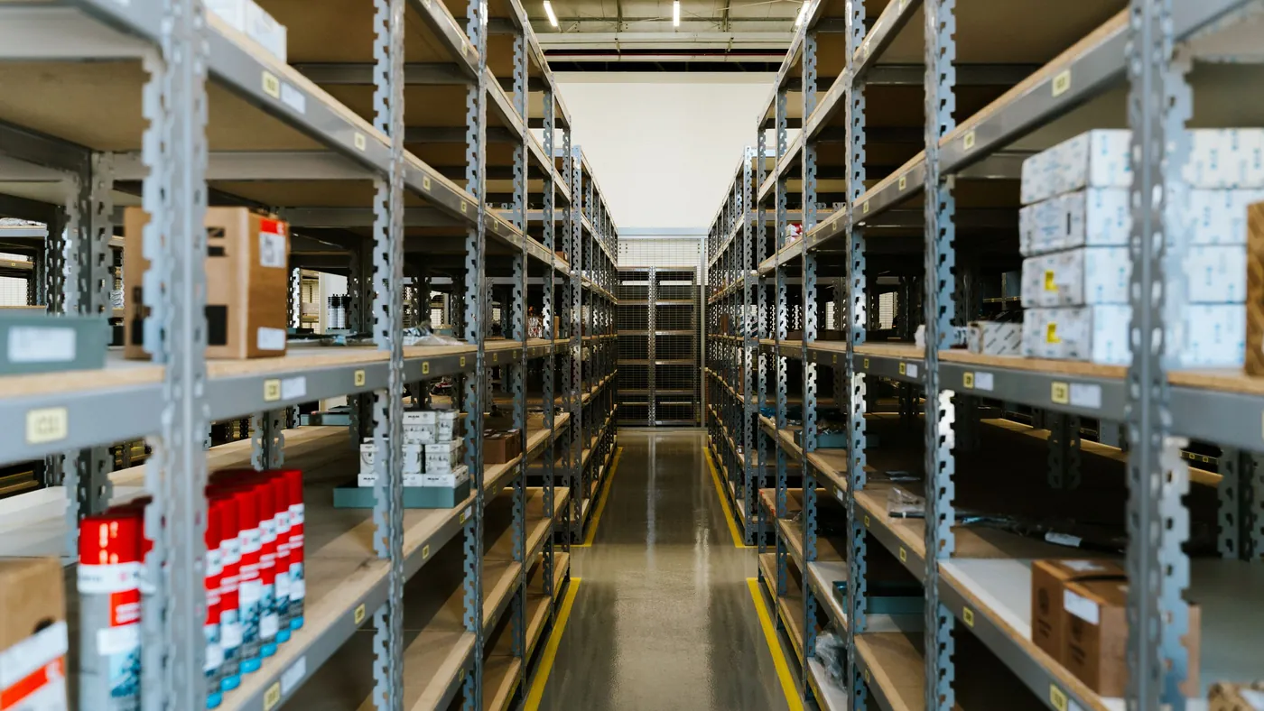 A long aisle of stocked metal warehouse shelves receding into the distance, standing in for a full catalog of real products that an AI agent still can't read.