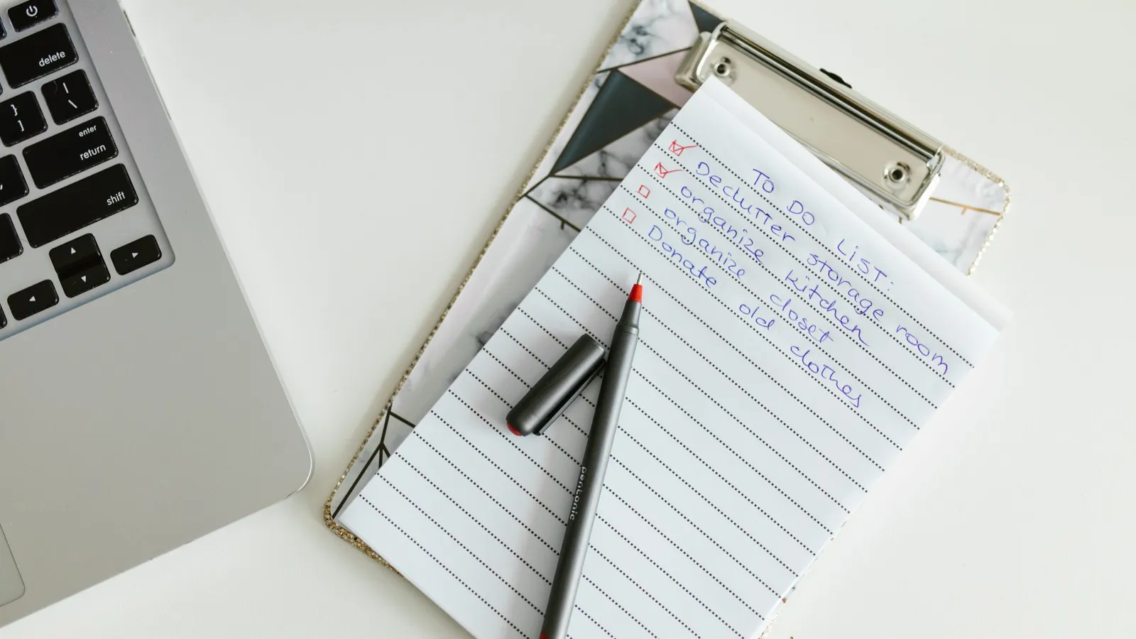 A handwritten to-do list on a clipboard beside a laptop keyboard, with two items ticked in red and a pen resting across the page.