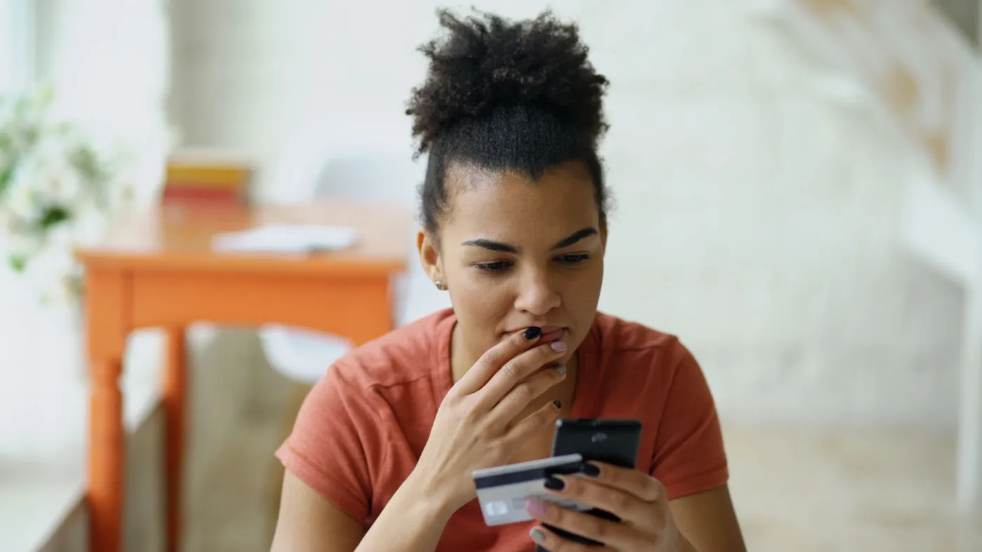 A young woman holds a bank card and a smartphone and pauses with her hand to her lips before confirming a purchase.
