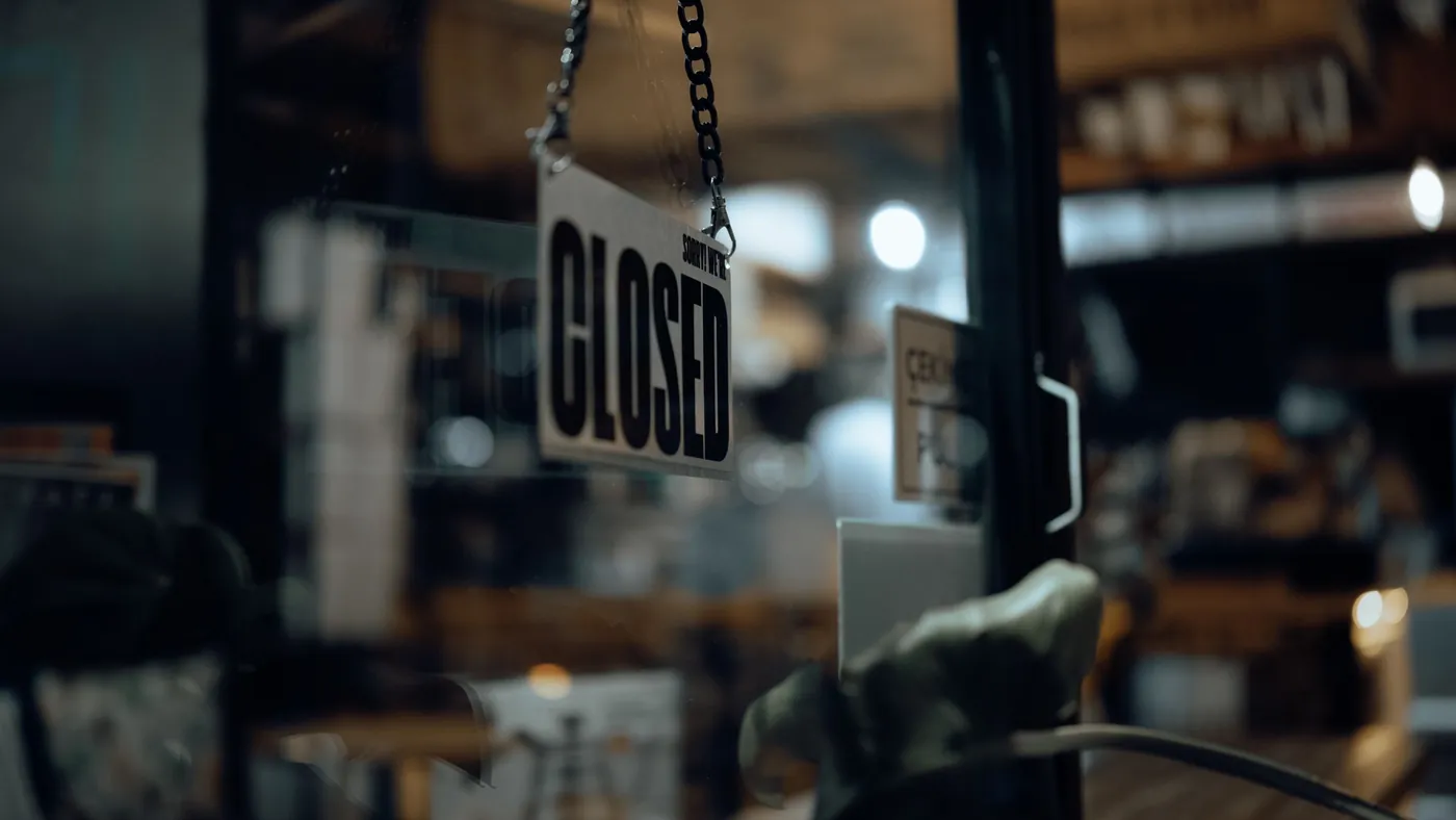 A closed sign hanging on a shop's glass door at night with the lit interior visible behind it, standing in for a store an AI agent can read in full detail but cannot complete a purchase at.