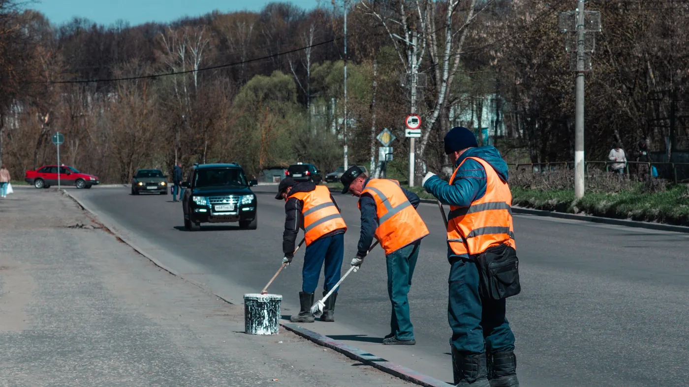 Three road workers in orange high-visibility vests repainting the white line along the edge of a road while traffic passes behind them.