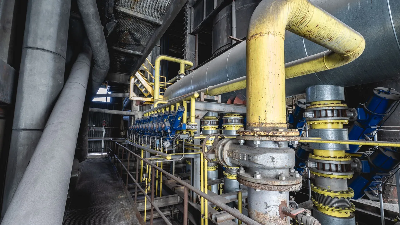 A dense industrial pipe run with a long row of blue valves and yellow pipework inside a large plant.