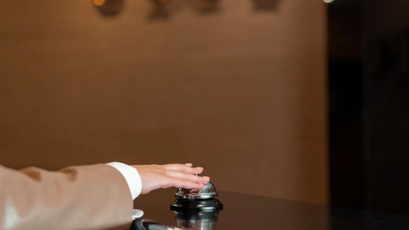 A hand pressing a silver service bell on a dark hotel reception counter, standing in for the booking and ordering systems that AI agents are now arriving at in food and lodging.