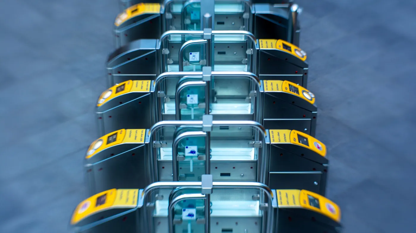 An overhead view of a row of ticket gates at a transit station, each one open but requiring a valid pass, standing in for controlling what an AI agent may do rather than trying to keep it out.